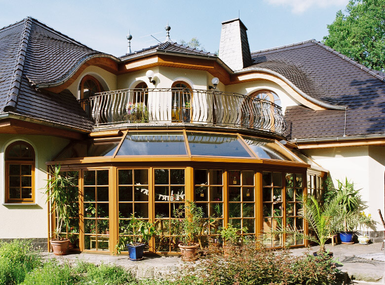 Der Eichenholz-Wintergarten unseres größten Massivhaus Grunewald dominiert das Winkelhaus. Das Biber-Walmdach ist geprägt von runden Ochsenaugen-Gauben. Die runden Sprossen-Holzfenstern und das braune Holz-Gesims imitieren bayerischen Villen-Hausbaustil. 